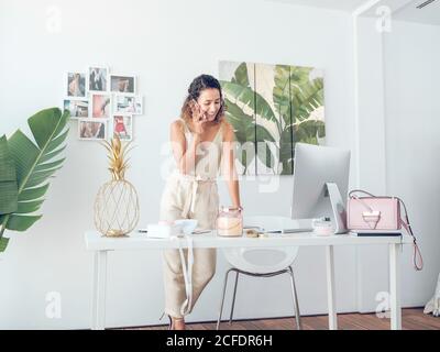 Bella donna in elegante vestito sorridente e di avere conversazione telefonica mentre si trova vicino alla scrivania in elegante ufficio Foto Stock