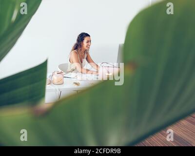 Donna giovane cute in elegante vestito sorridente e digitando sulla tastiera del computer mentre seduto alla scrivania in elegante ufficio leggero Foto Stock