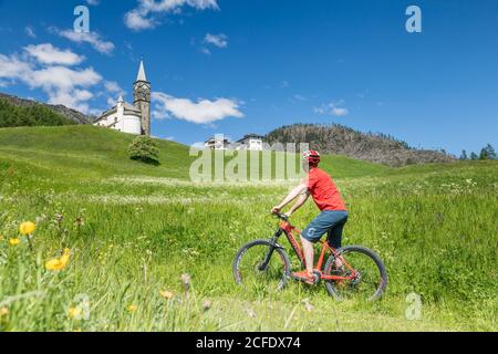 ciclista in e-bike a laste di rocca pietore, nei prati di fronte alla chiesa parrocchiale, dolomiti, agordino, belluno, veneto, italia Foto Stock