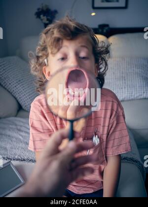 Bambino curly sorpreso che mostra dente con la bocca larga tirare abbassare il labbro mentre la persona tiene la lente d'ingrandimento nella stanza Foto Stock