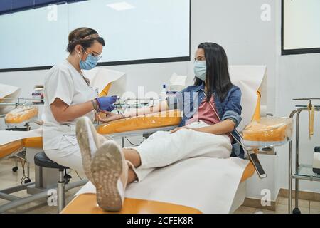 Vista laterale della giovane donna in maschera protettiva seduta in poltrona medica durante la procedura di trasfusione di sangue in ospedale contemporaneo Foto Stock