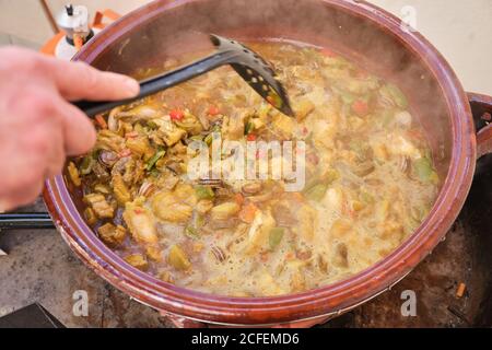 Crop anonimo uomo in piedi che puttano skimmer in grande pentola di argilla con piatto da cucina di riso di pollo verdure assortite e lumache Foto Stock