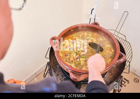 Crop anonimo uomo in piedi che puttano skimmer in grande pentola di argilla con piatto da cucina di riso di pollo verdure assortite e lumache Foto Stock