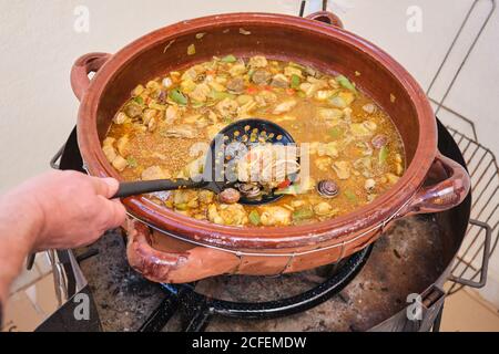 Crop anonimo uomo in piedi che puttano skimmer in grande pentola di argilla con piatto da cucina di riso di pollo verdure assortite e lumache Foto Stock