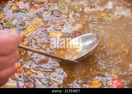 Crop anonimo uomo in piedi che puttano skimmer in grande pentola di argilla con piatto da cucina di riso di pollo verdure assortite e lumache Foto Stock