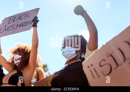 Donne etniche serie con acconciatura afro che tiene poster di cartone con black lives materia iscrizione e alzando pugno durante la dimostrazione in città affollata e aggrapparsi Foto Stock