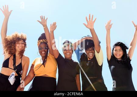 Basso angolo di compagnia di donne multietniche amichevoli che esprimono gioia con braccia sollevate sullo sfondo del cielo senza nuvole in estate e guardando la fotocamera Foto Stock