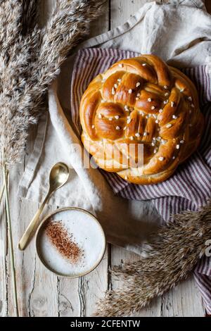 Tazza di cappuccino caldo seduto al tavolo di legno con pane fresco fatto in casa intrecciato posto su tessuto accanto all'animale pelliccia Foto Stock