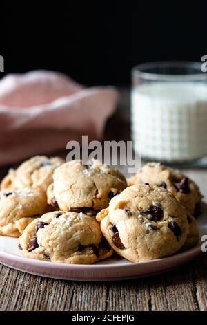 Piatto di gustosi biscotti vegani con gocce di cioccolato tavolo di legno vicino al latte sfocato e panno su sfondo nero Foto Stock