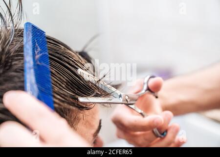 Vista laterale di maschio irriconoscibile che fa un taglio di capelli a ragazzo usando forbici contro gli interni sfocati del bagno leggero a casa Foto Stock