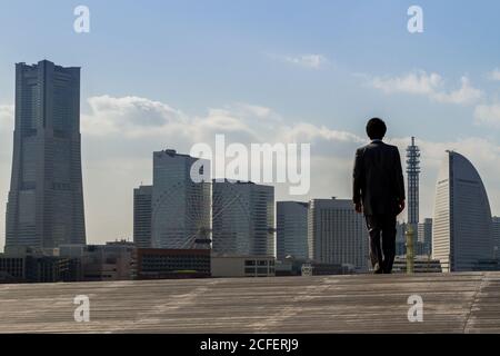 Un salarista giapponese sorge sul Molo di Osanbashi guardando lo skyline di Minato Mirai con la Landmark Tower (a sinistra) Yokohama. Kanagawa Giappone. Foto Stock