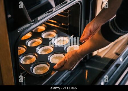 Alto angolo di casalinga raccolto mettendo vassoio di muffin con l'impasto in casse di carta in forno mentre cucinando in cucina Foto Stock