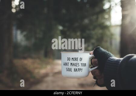 Mano del viaggiatore anonimo che tiene la tazza di metallo con citazione ispiratrice sullo sfondo sfocato della foresta in giornata di sole Foto Stock