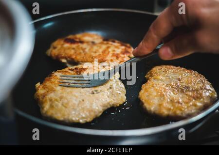 Closeup di mano di raccolto di persona anonima che cucinano carne deliziosa le cotolette frittura sull'olio sulla padella calda in cucina Foto Stock