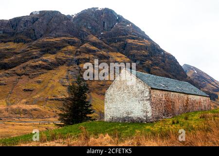 Casa in pietra invecchiata situata su una collina verde contro maestose rocce montagna nelle Highlands scozzesi Foto Stock