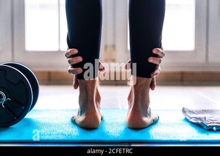 Vista posteriore di un uomo a piedi nudi flessibile irriconoscibile in abbigliamento sportivo fare in piedi avanti curva esercizio vicino dumbbell mentre allenarsi da solo tappetino sportivo contro la finestra in una stanza luminosa e spaziosa a casa Foto Stock