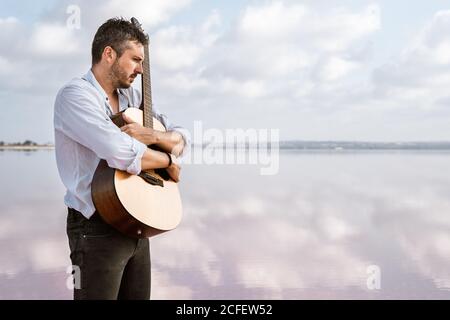 Uomo appassionato malinconico in camicia bianca e sospenditori che abbracciano la chitarra mentre si è in piedi nudi in acqua a riva in giorno nuvoloso Foto Stock