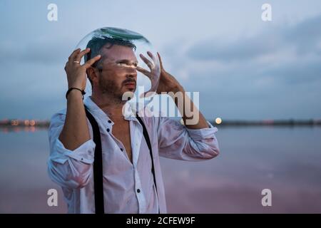 Uomo pusivo in camicia bagnata con acquario vuoto sulla testa stare in piedi e contemplare dal mare fermo al crepuscolo Foto Stock