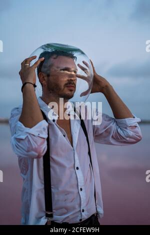 Uomo pusivo in camicia bagnata con acquario vuoto sulla testa stare in piedi e contemplare dal mare fermo al crepuscolo Foto Stock