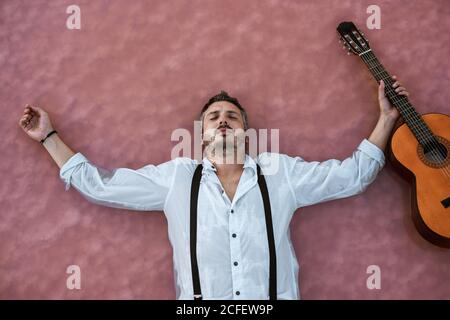 Vista dall'alto di un uomo assonnato in camicia bianca e sospetti sdraiato sulla schiena con gli occhi chiusi e fluttuante con acustica chitarra in mare a sandbank Foto Stock