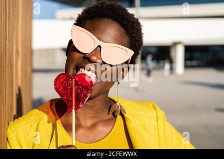 Donna afroamericana alla moda in occhiali da sole in giacca gialla godendo a forma di cuore lollipop da recinzione di legno Foto Stock