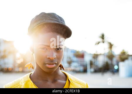 Ritratto di Afro American Woman in elegante giacca luminosa guardando la fotocamera su spiaggia sabbiosa sfondo sfocato Foto Stock