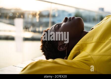 Vista dall'alto dell'elegante donna afroamericana nera in giacca moderna rilassante su panca di legno e guardando lontano Foto Stock