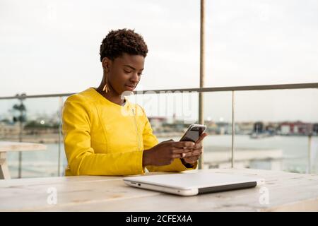 Concentrato afroamericano donna di messaggistica smartphone mentre rilassarsi a legno tabella Foto Stock