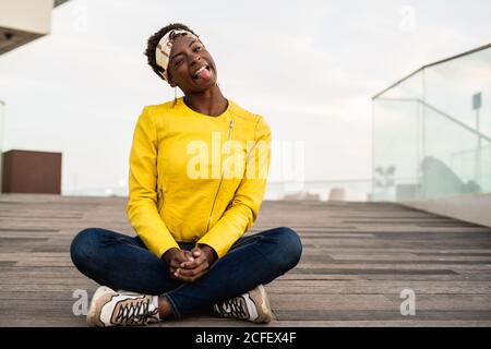 Elegante donna afroamericana in giacca moderna rilassante seduta su pavimento di legno e guardando la macchina fotografica che fa i volti sciocco Foto Stock