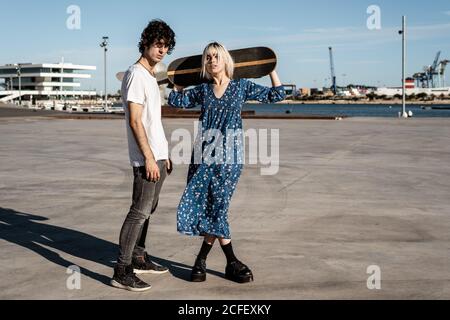 Giovane coppia affettuosa alla moda in piedi mentre la donna tiene uno skateboard in piazza contro il cielo blu e gli edifici moderni sfocati in condizioni di vento Foto Stock