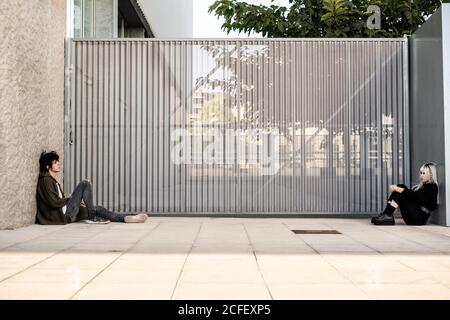 Vista laterale dell'infelice fiero Donna sola e dell'uomo seduti l'uno di fronte all'altro sul terreno appoggiato sul muro e contemplando dopo il disaccordo durante la crisi del rapporto vicino al cancello dell'edificio moderno sulla strada Foto Stock