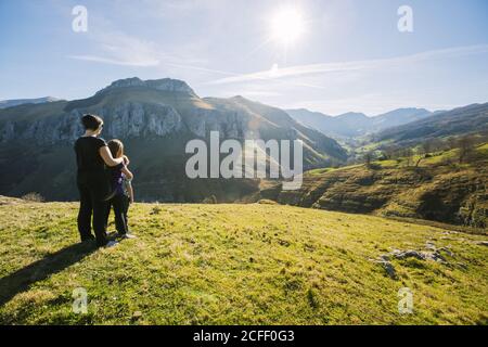 Vista posteriore di hiker femminile anonimo abbracciare i bambini mentre si è in piedi sul prato verde e osservando il paesaggio pittoresco delle montagne sopra Giornata di sole in Cantabria Foto Stock