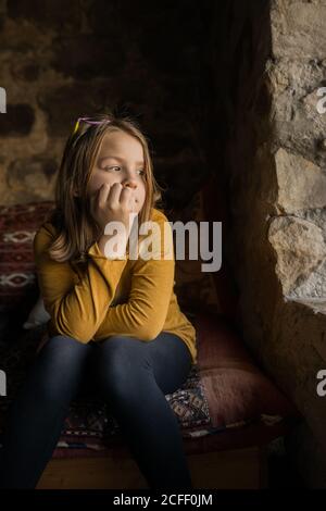 Ragazza pensierosa solitaria in vestiti casual appoggiata a mano mentre seduto sul divano e guardando lontano nella finestra di vecchio Casa in pietra in Spagna Foto Stock