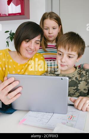 Donna adulta e bambini che navigano in un tablet moderno insieme mentre sono seduti al tavolo a casa Foto Stock