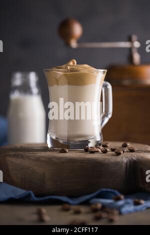 Caffè montato caffè Dalgona nel latte freddo posto vicino ai chicchi di caffè, macinacaffè e bottiglia di latte sulla superficie rustica di legno rotondo con tovagliolo blu sotto Foto Stock