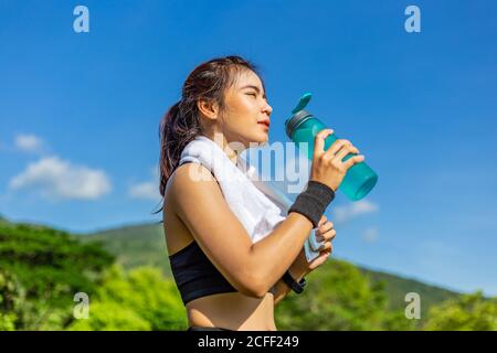 Bella giovane donna asiatica che si esercita al mattino su una pista di corsa prendendo un riposo per bere acqua, profondo sfondo blu cielo in giornata di sole luminoso Foto Stock