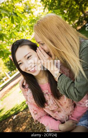 Giovane donna caucasica sorridente e sussurrante segreto in orecchio di sorridente amico asiatico mentre si passa il tempo in parco insieme Foto Stock