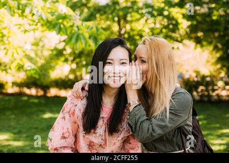 Giovane donna caucasica sorridente e sussurrante segreto in orecchio di sorridente amico asiatico mentre si passa il tempo in parco insieme Foto Stock