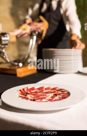 Crop persona irriconoscibile mano sul grembiule tagliando un intero secco-indurito prosciutto gamba su sfondo nero Foto Stock