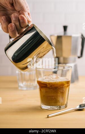 Barista professionista senza volto che versa il latte da una caraffa in metallo in vetro con caffe' fresco su tavolo di legno nel cafe' Foto Stock