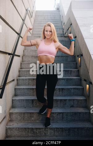 Giovane bionda caucasica Donna che flette i muscoli con abbigliamento sportivo all'aperto Foto Stock