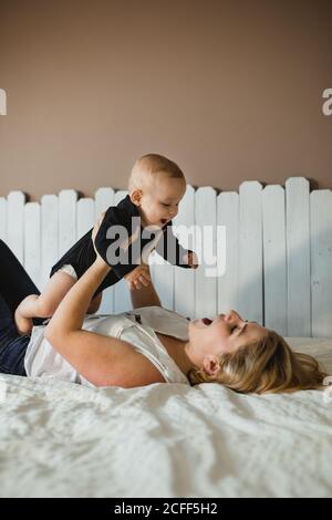 Vista laterale di giovane donna eccitato che solleva il bambino carino e gioca mentre sdraiato sul letto comodo a casa Foto Stock