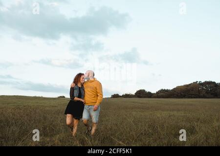 Giovane coppia allegra e romantica che tiene le mani e cammina sul verde campo al mare al tramonto con cielo nuvoloso Foto Stock