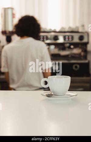 Tazza bianca di caffè con piatto e cucchiaio banco bar con barista femminile sfocato che utilizza una macchina da caffè in background Foto Stock