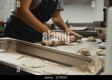 Vista laterale del panettiere prodotto in grembiule nero che si arrotola impastare sul tavolo con la farina bianca mentre si fa il pane piatto in cucina Foto Stock