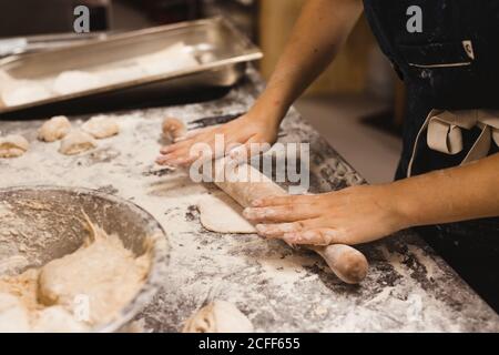 Vista laterale del panettiere prodotto in grembiule nero che si arrotola impastare sul tavolo con la farina bianca mentre si fa il pane piatto in cucina Foto Stock