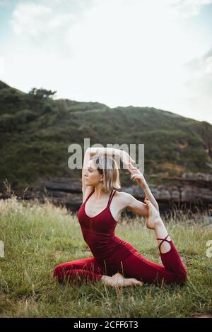 Giovane donna flessibile in abbigliamento sportivo rosso seduta in yoga difficile uno zampe king piccione posa su collina verde vuoto Foto Stock