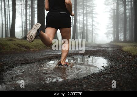 Vista posteriore di una corridore femminile senza volto nell'allenamento activewear in boschi e correre attraverso puddle sporco in boschi Foto Stock