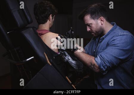 Master facendo tatuaggio sull'avambraccio del cliente maschile Foto Stock