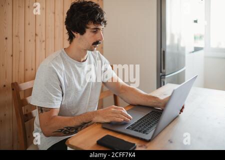 Vista laterale focalizzato freelance maschile seduto al tavolo in cucina e lavorando al progetto mentre si utilizza il computer Foto Stock